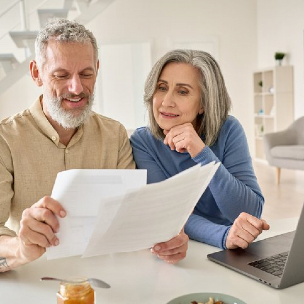 Middle aged senior old couple holding estate planning documents and using laptop at home.