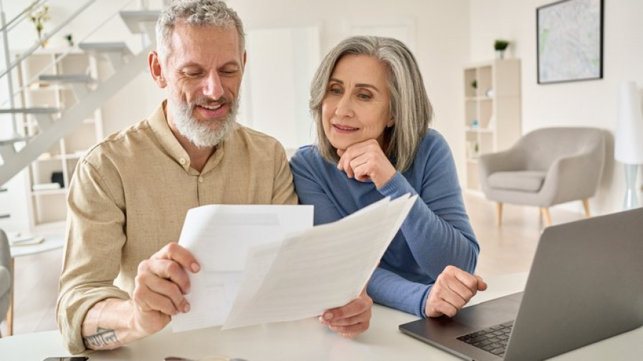 Middle aged senior old couple holding estate planning documents and using laptop at home.