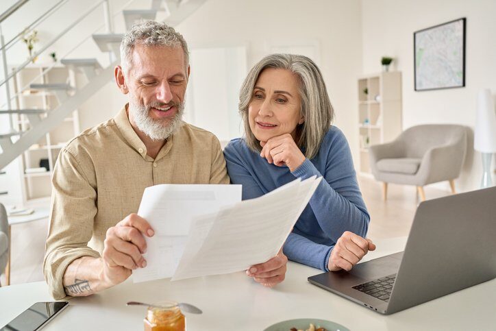 Middle aged senior old couple holding estate planning documents and using laptop at home.